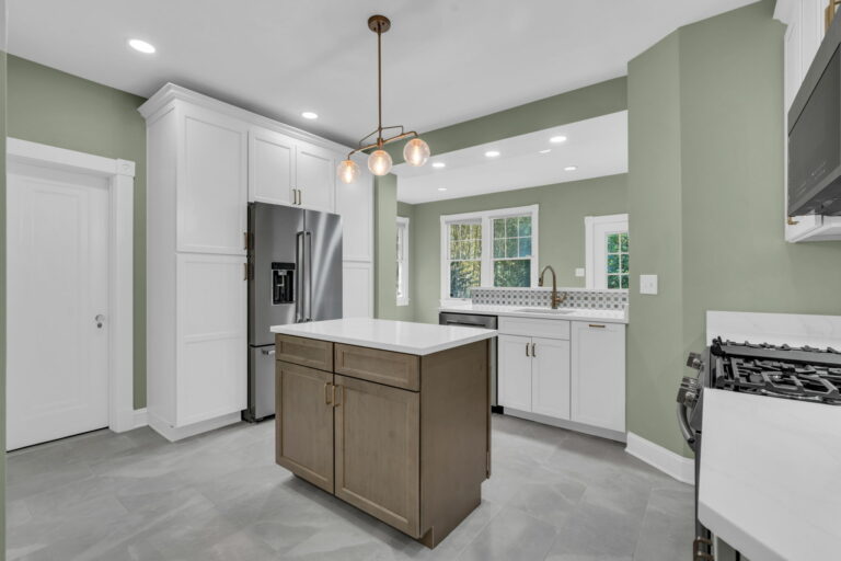 This Silver Spring kitchen remodel by Mega Design and Build combines contemporary style with practical design for everyday living and entertaining. Crisp white cabinetry contrasts beautifully with calming sage walls, creating an airy and welcoming atmosphere. Durable quartz countertops grace the central wood-tone island, which features both storage and bar seating. Modern appliances, including a French door refrigerator and gas range, are complemented by a geometric tile backsplash. Recessed and pendant lighting highlights the space perfectly. Wide windows bring in natural light and enhance the kitchen’s openness.