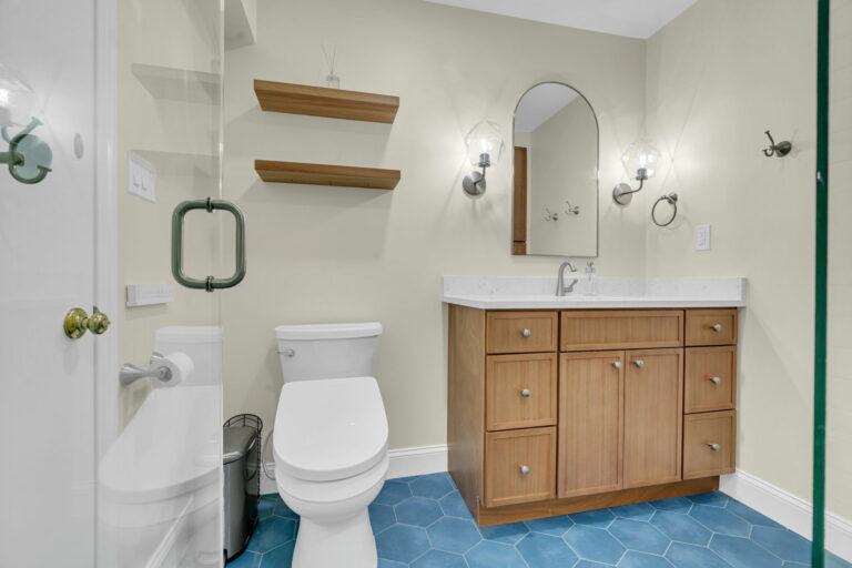 A rainfall showerhead and bold blue-gray hexagonal floor tiles add a spa-like touch to the bright, subway-tiled shower area. Warm wood vanities contrast beautifully with the crisp white marble countertops and the central mirror. Space-saving floating shelves, towel hooks, robe rings, and task lighting ensure everyday convenience. The thoughtful mix of textures and neutral colors, enhanced by standout tiles, creates a modern bathroom that is both stylish and efficient.