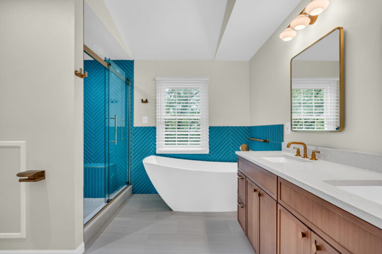 The crisp white surroundings create a balanced backdrop for this vibrant focal point, while large windows flood the space with natural light. Warm-toned wood vanities complement the gray tiled floor, and shimmering gold fixtures, shower hardware, and framed mirrors add a touch of luxury. The glass-enclosed shower keeps the space feeling open, and recessed and sconce lights provide thoughtful illumination.